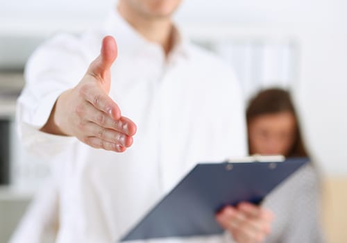 a person holds out a hand to shake while holding a clipboard while one of several clients sits behind them